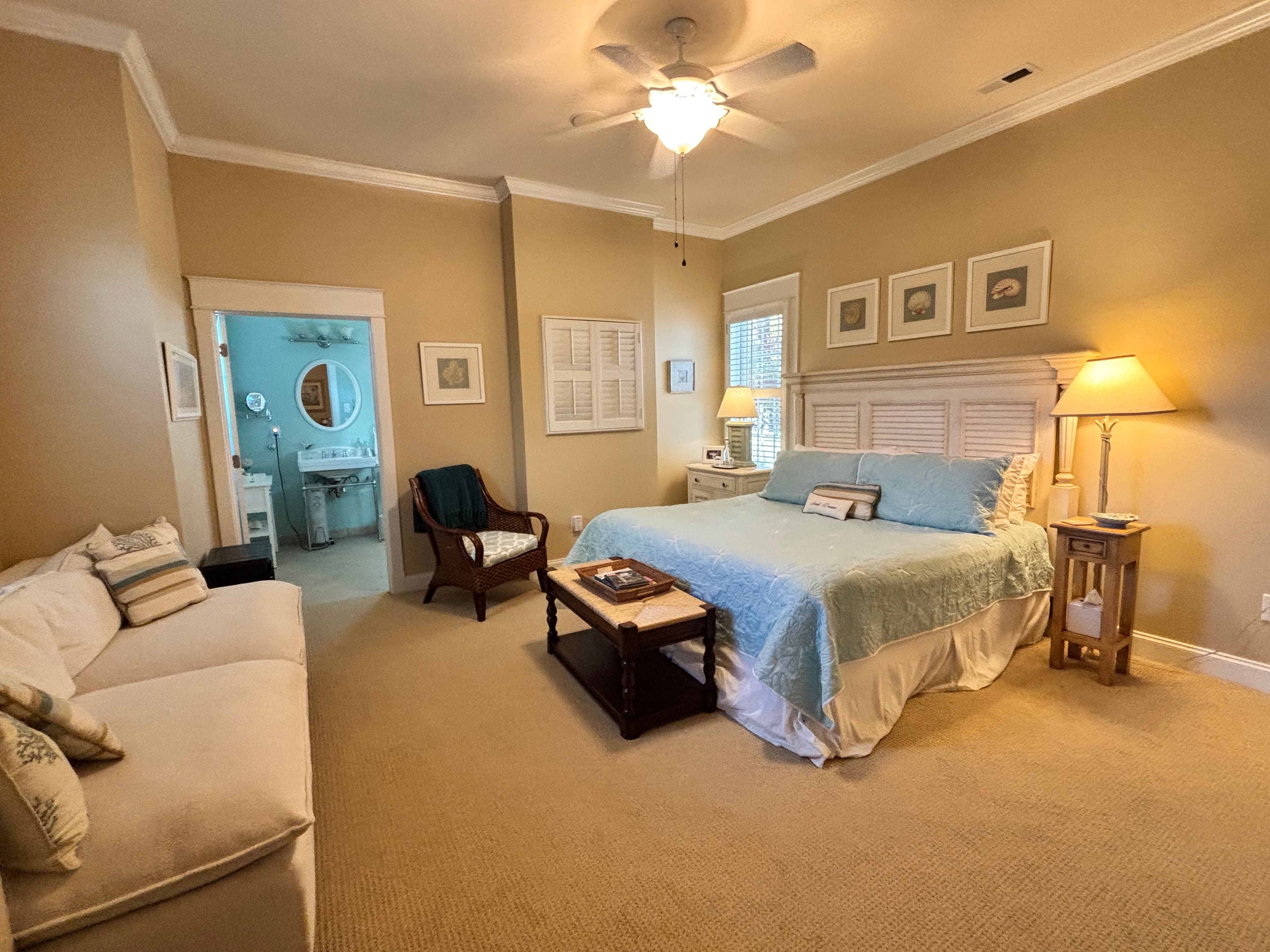Inviting guest suite with coastal décor featuring a king bed with light blue bedding, cozy sitting area, and en-suite bathroom. The room includes soft beige walls, beach-inspired artwork, and natural lighting - perfect for a relaxing getaway or bed and breakfast stay.