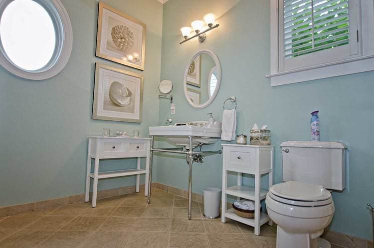 A modern bathroom with light blue walls, a round mirror, decorative artwork, and white fixtures.