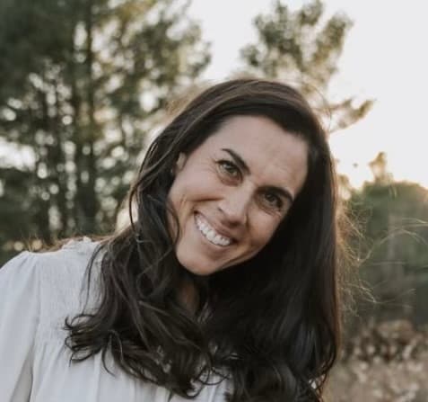 A woman with long dark hair smiles warmly in a sunlit outdoor setting.