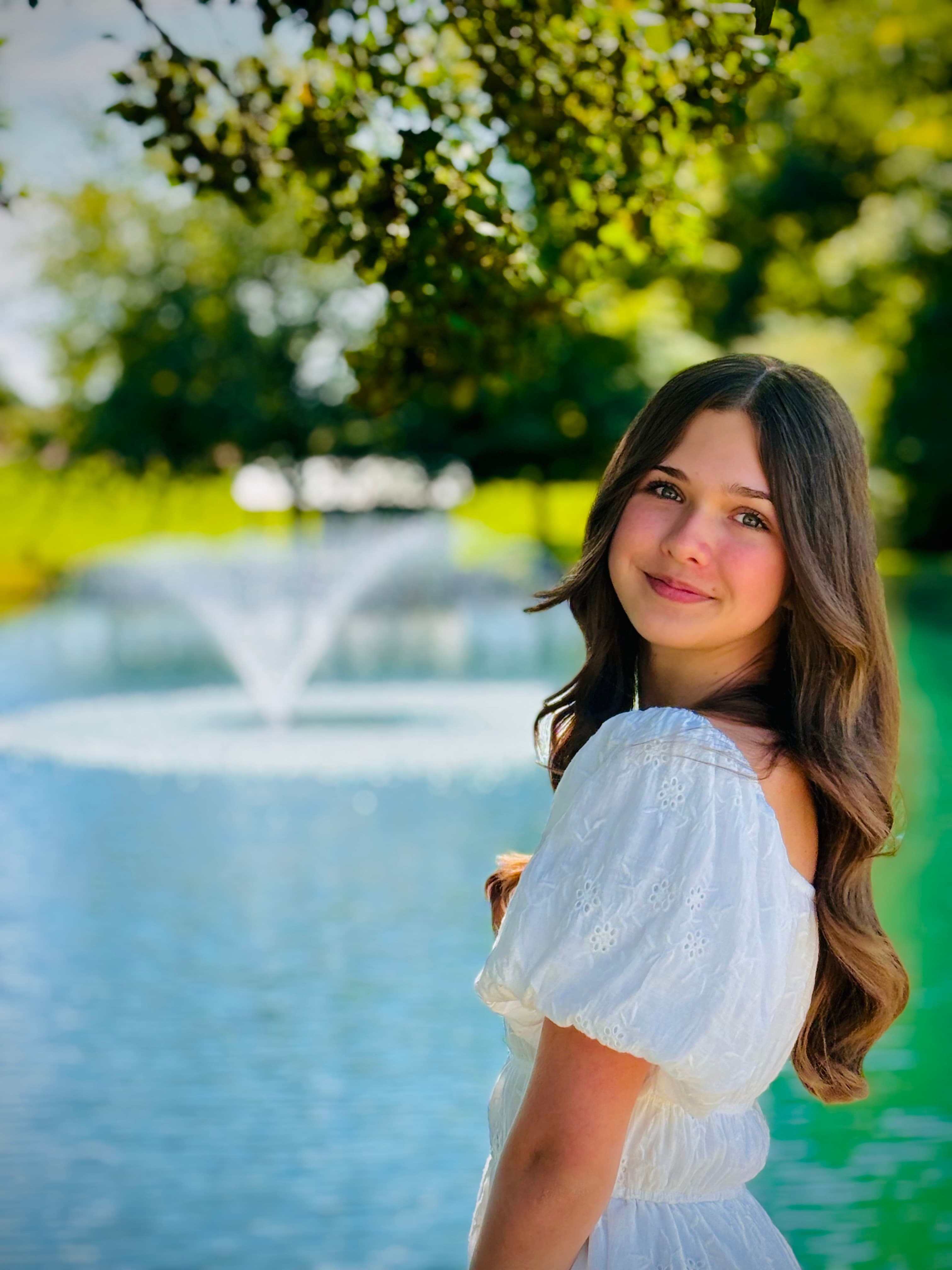 A young woman in a white dress smiles near a fountain in a serene park setting.