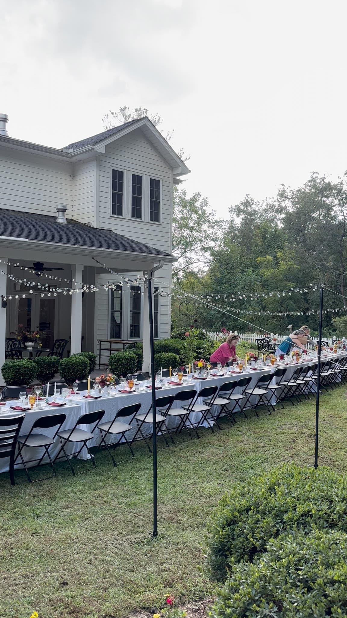 An outdoor table set for a gathering, adorned with flowers and surrounded by greenery, next to a house.