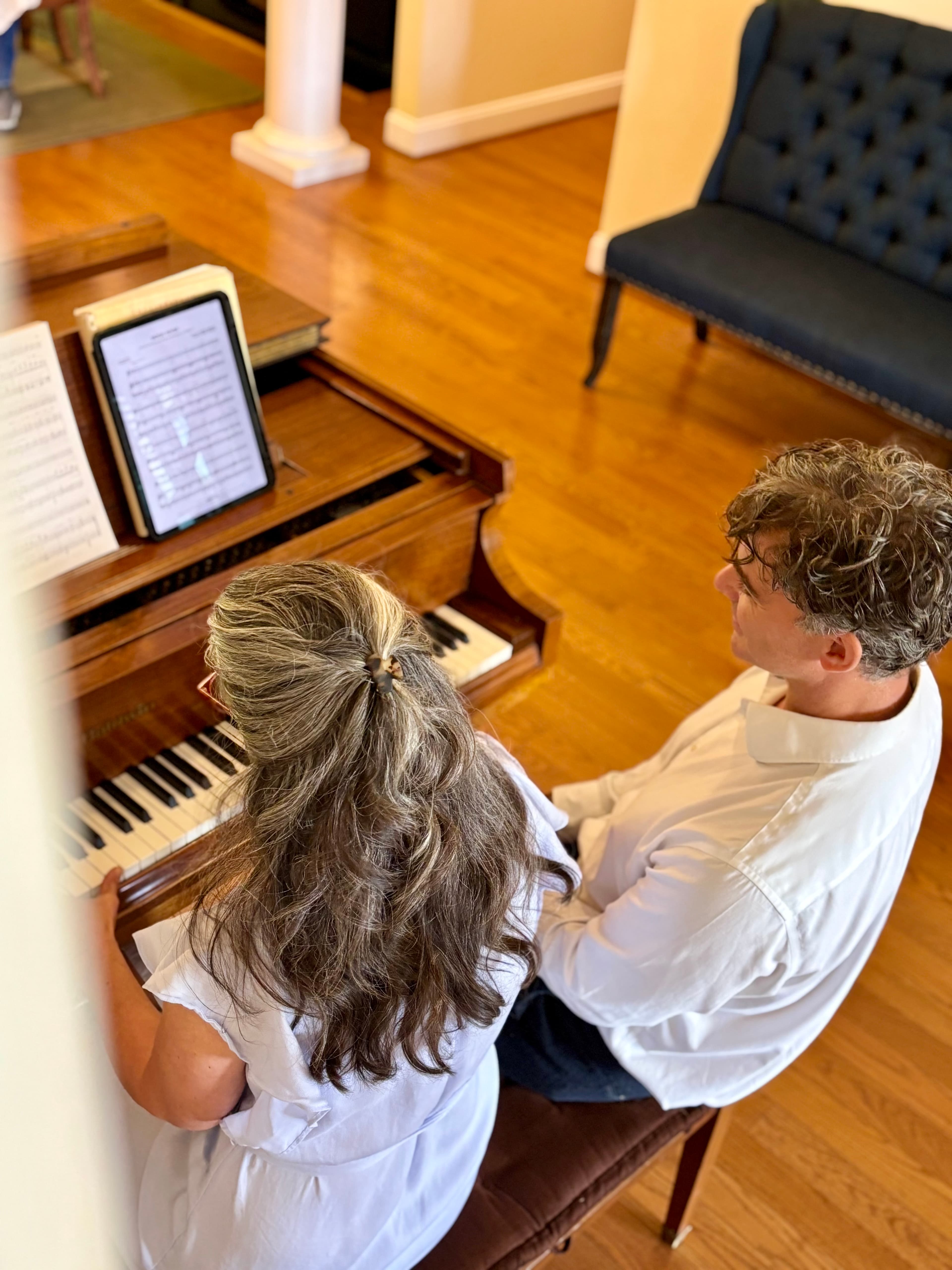 Two people, a woman and a man, sit together at a piano, reading music from a tablet.