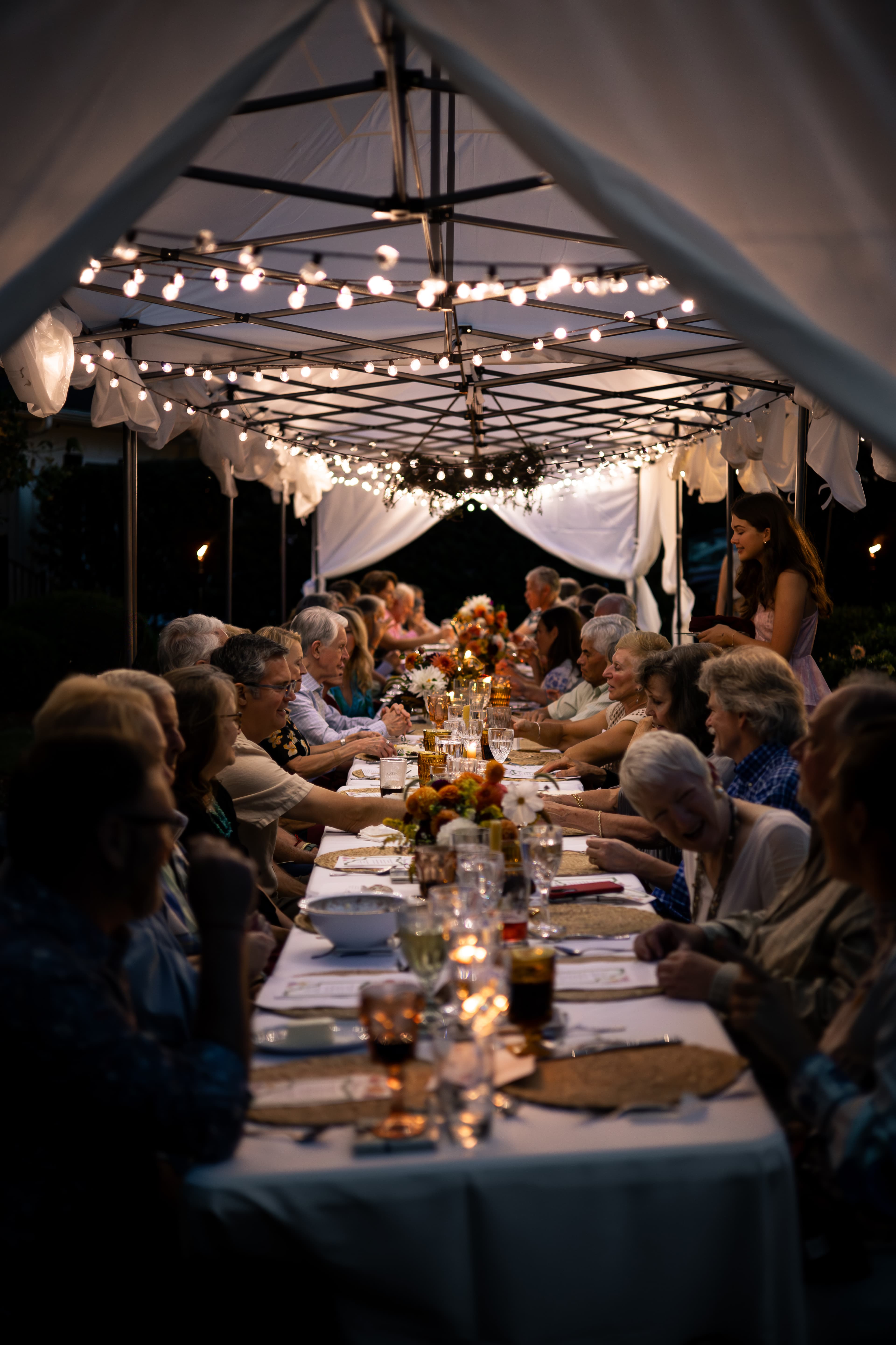 A long table filled with diners enjoying a meal under a tent adorned with string lights. A long table filled with diners enjoying a meal under a tent adorned with string lights.