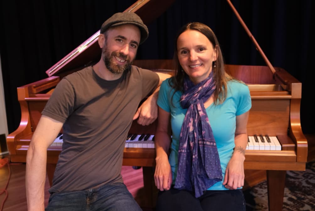 A man and a woman pose together in front of a grand piano, both smiling at the camera.