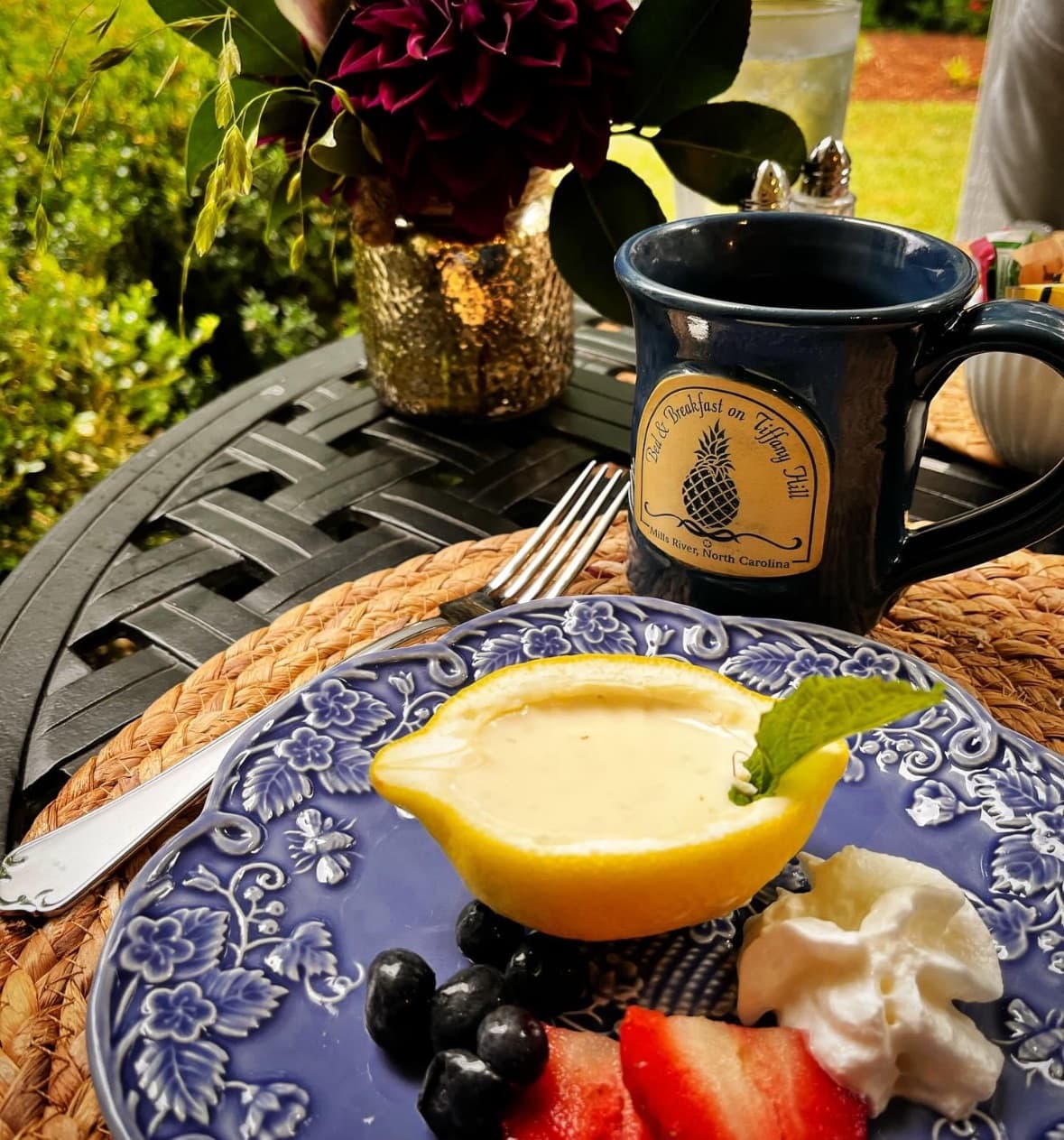 A blue plate holds a lemon dessert, berries, and whipped cream, alongside a coffee mug and a floral centerpiece on a patio table.