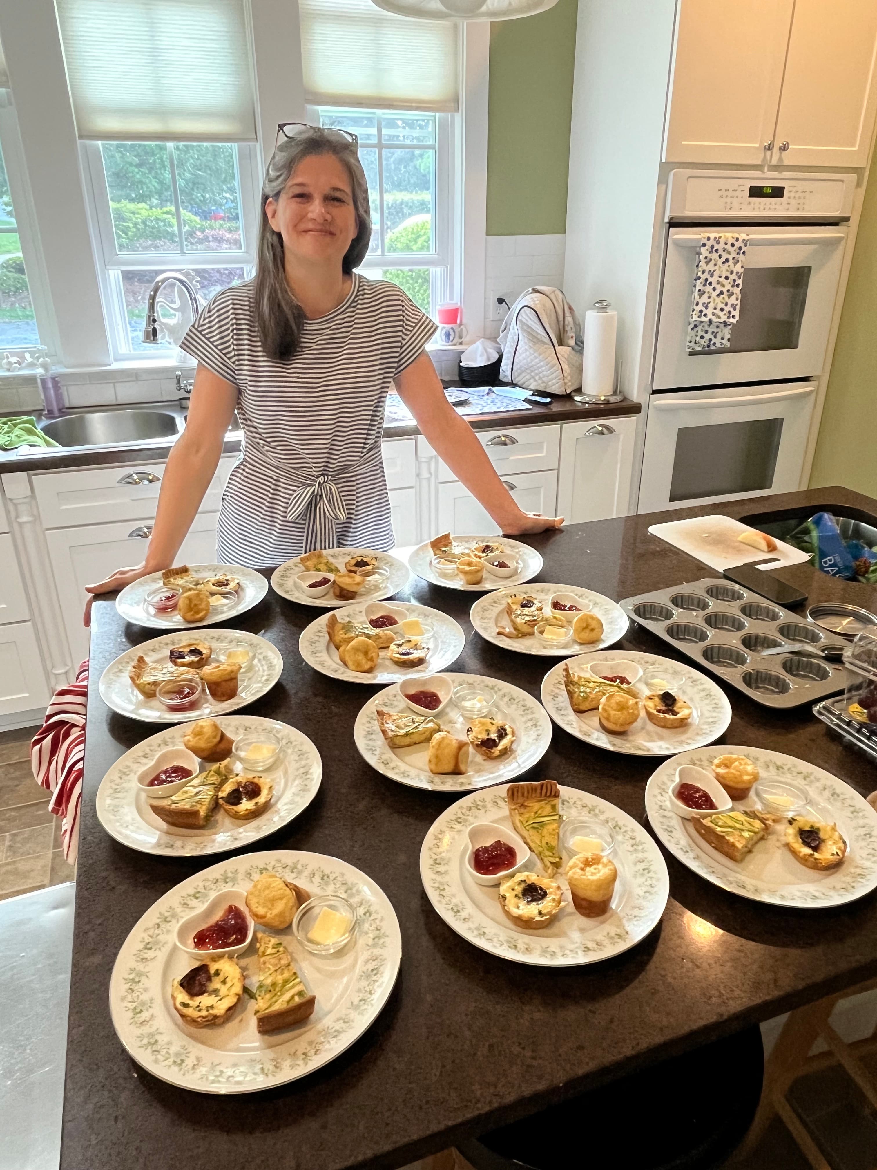 A woman stands smiling in a kitchen surrounded by plates of various baked goods.