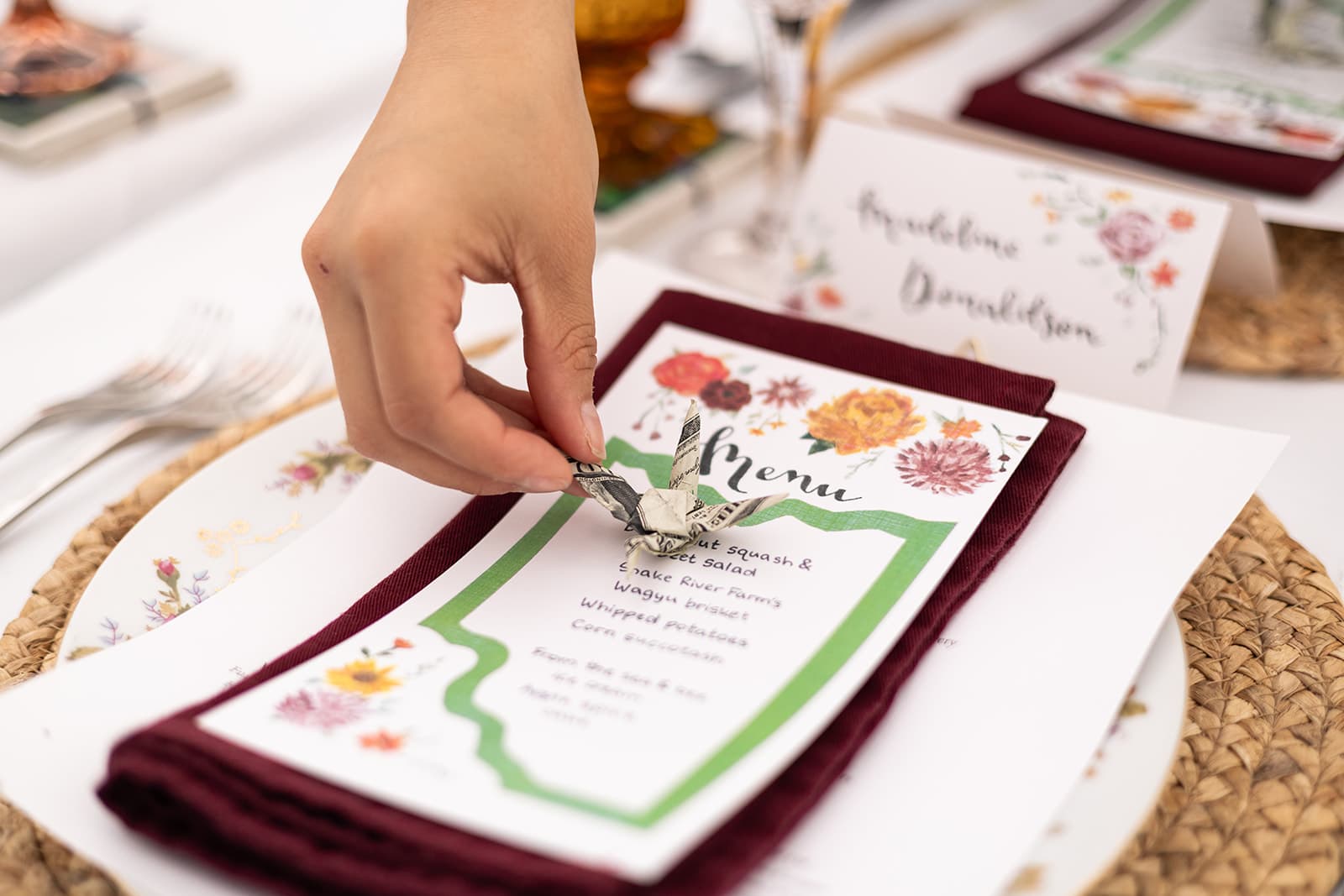 A hand is placing a decorative item on a menu card resting on a maroon napkin at a beautifully set dining table.