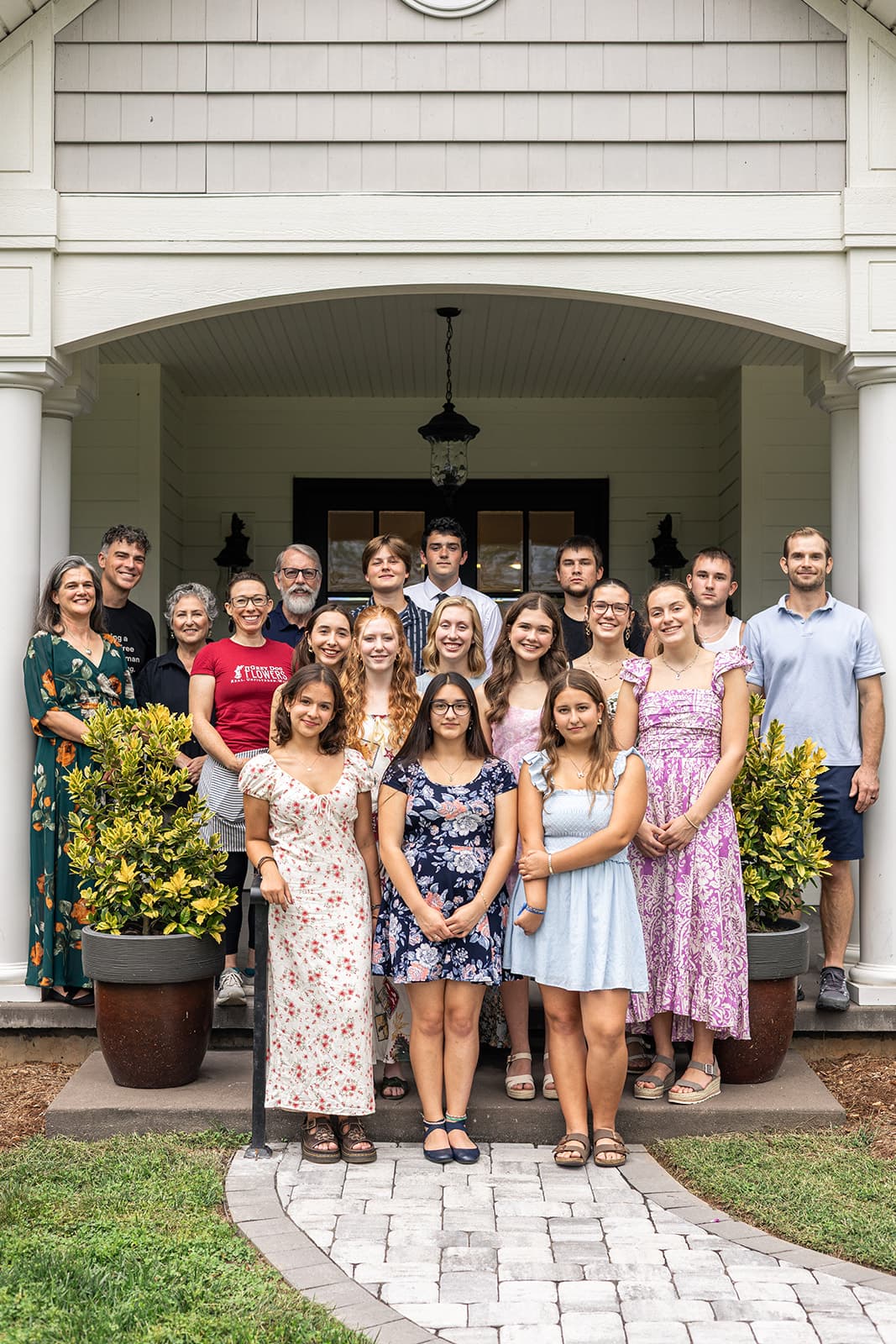A large group of people, including adults and teenagers, poses together on a porch surrounded by potted plants.