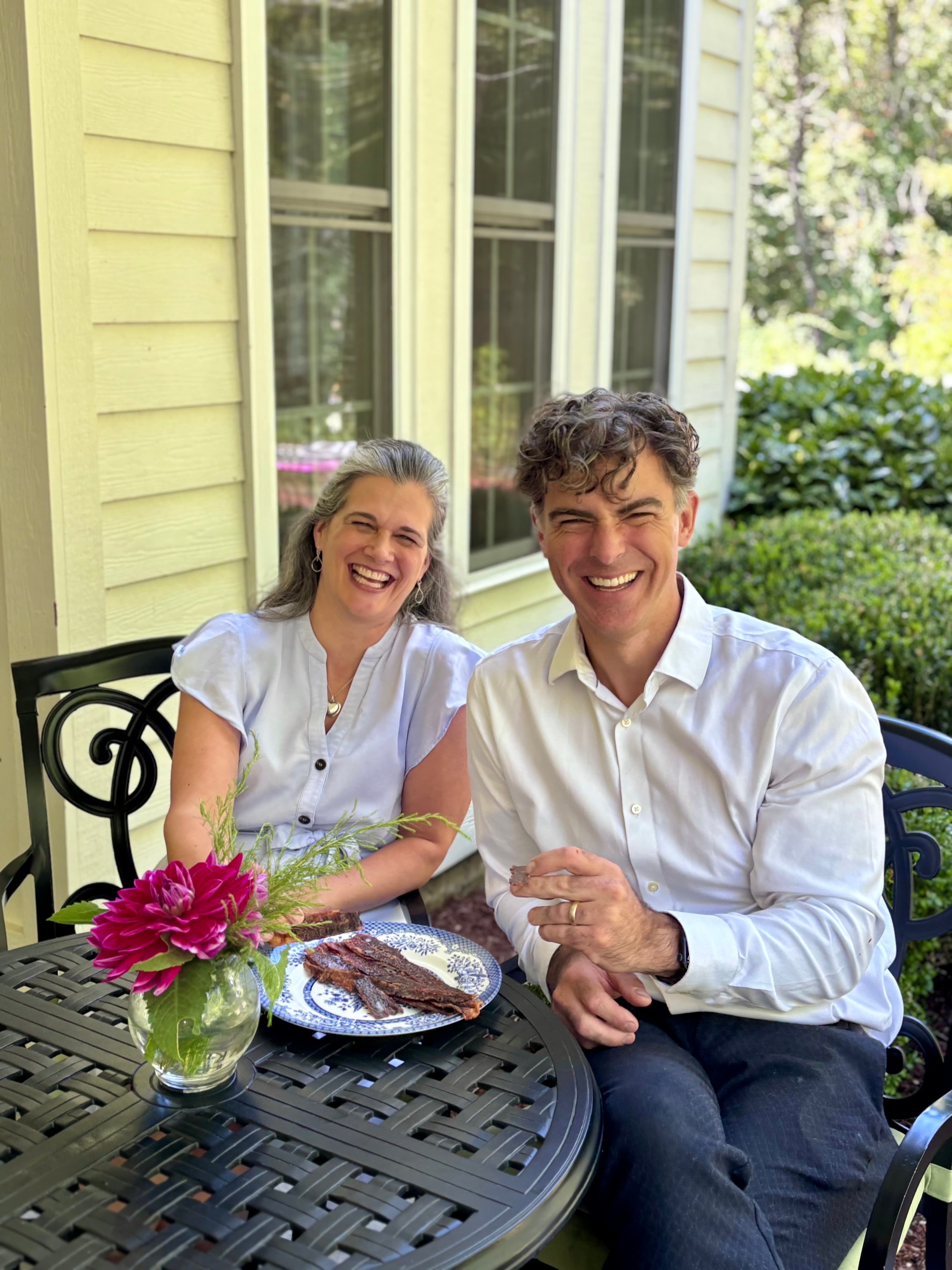 A man and woman smile joyfully while seated at a table outdoors, adorned with a small flower vase and a plate of food.