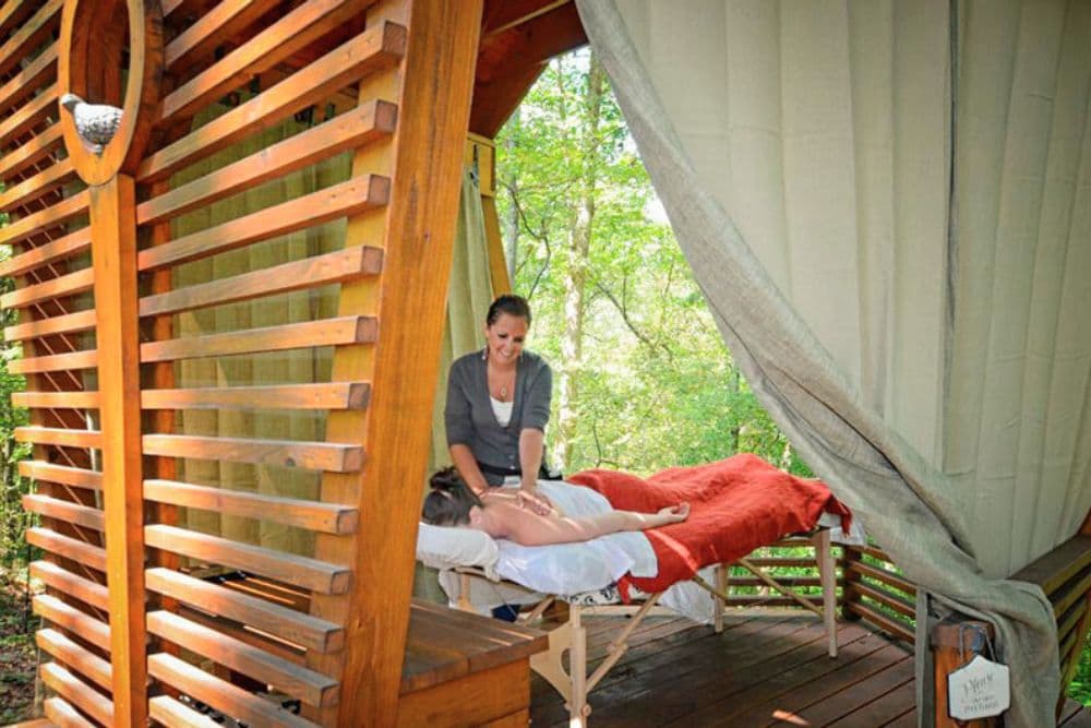 A massage therapist gives a massage to a client in a wooden outdoor setting surrounded by greenery.