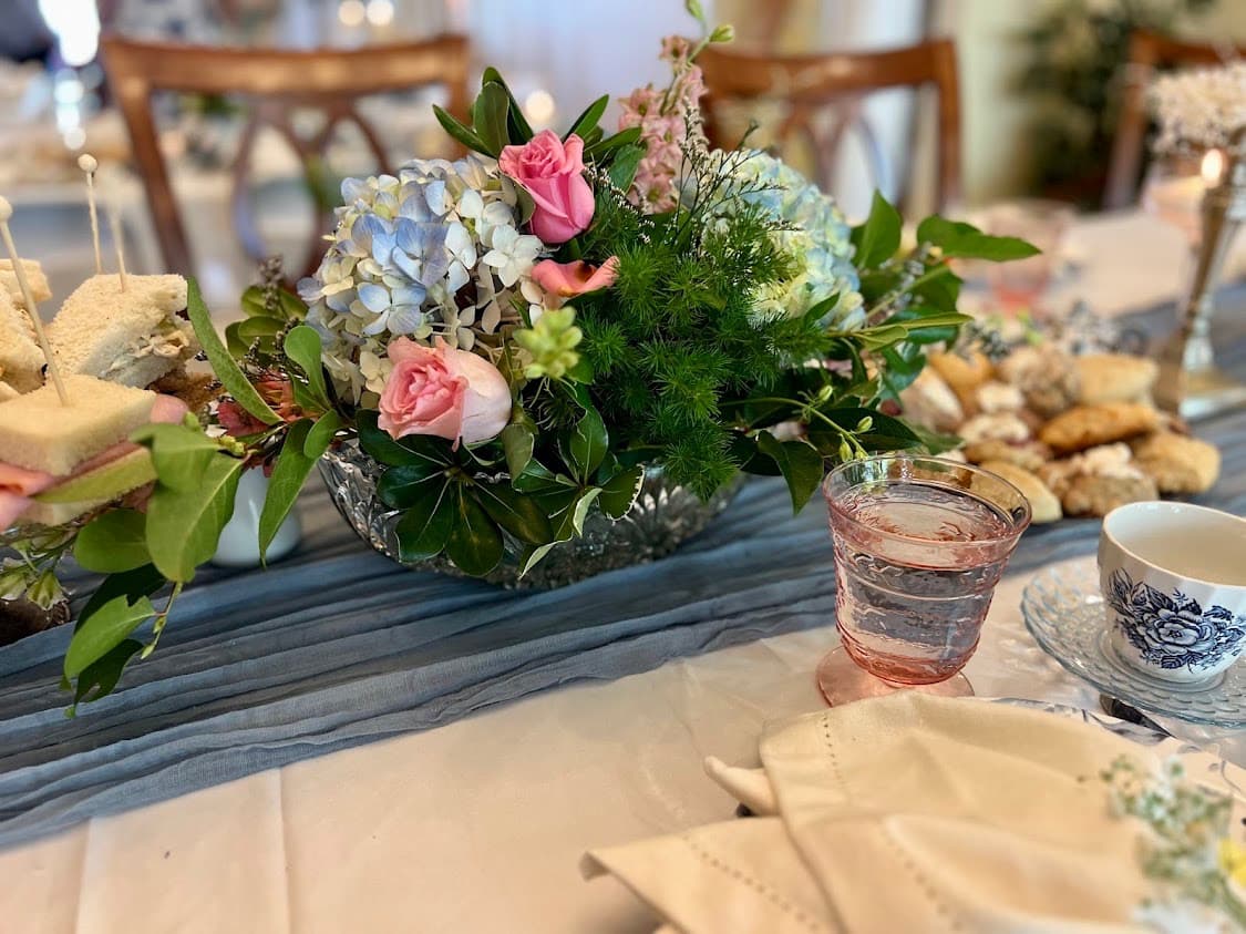 A beautifully arranged floral centerpiece with pink roses and hydrangeas beside tea, water, and assorted treats on a laid table.