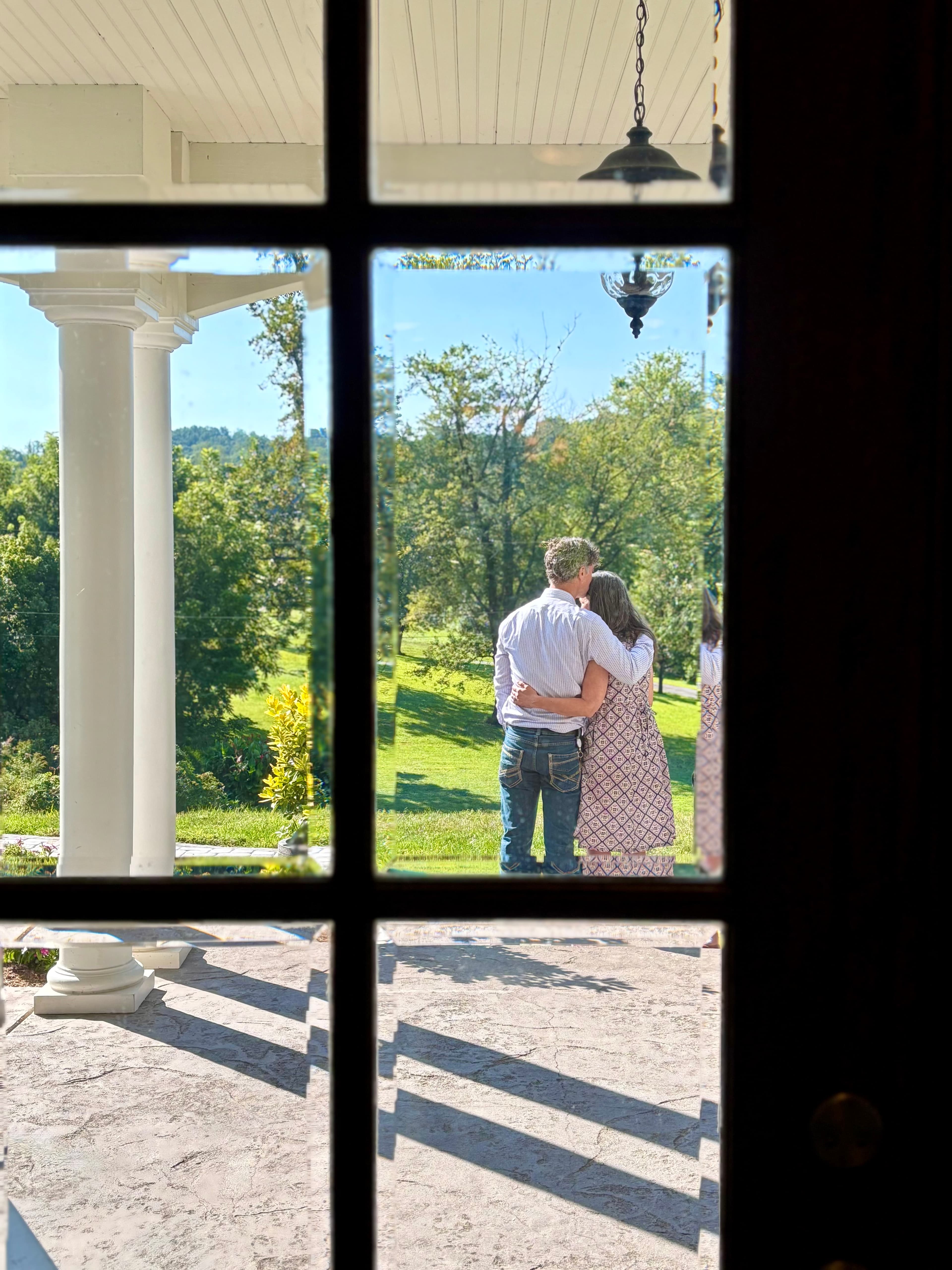 A couple embraces while looking out over a lush, green landscape through a window.