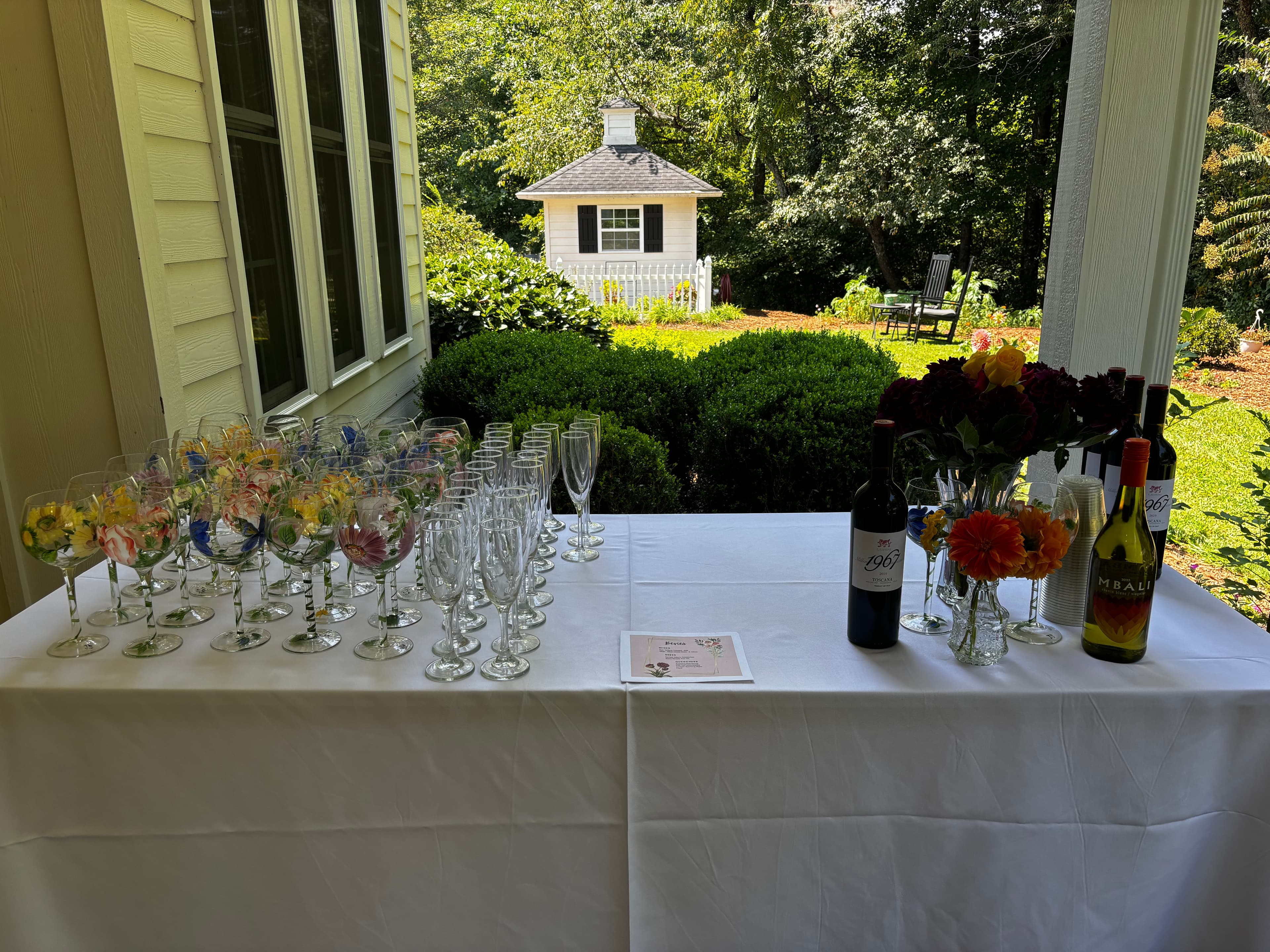 A table set with colorful wine glasses, champagne flutes, wine bottles, and a bouquet of flowers in an outdoor setting.