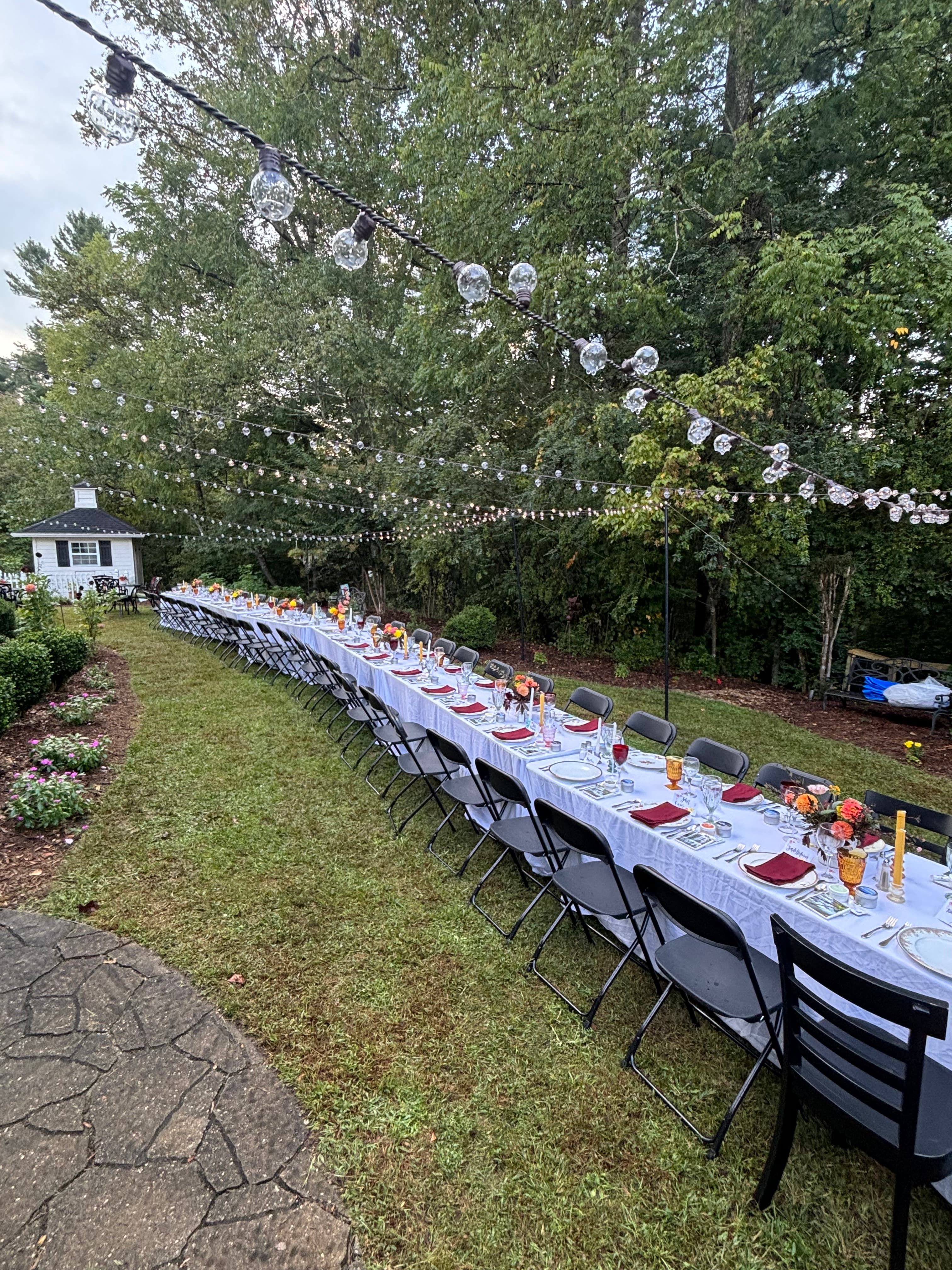 A long outdoor dining table is set with dishes and decorations under string lights surrounded by trees.
